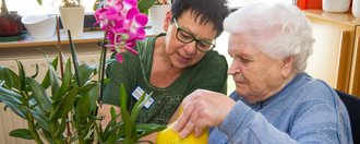 eine Pflegerin hilft einer Frau beim Gießen einer Orchidee