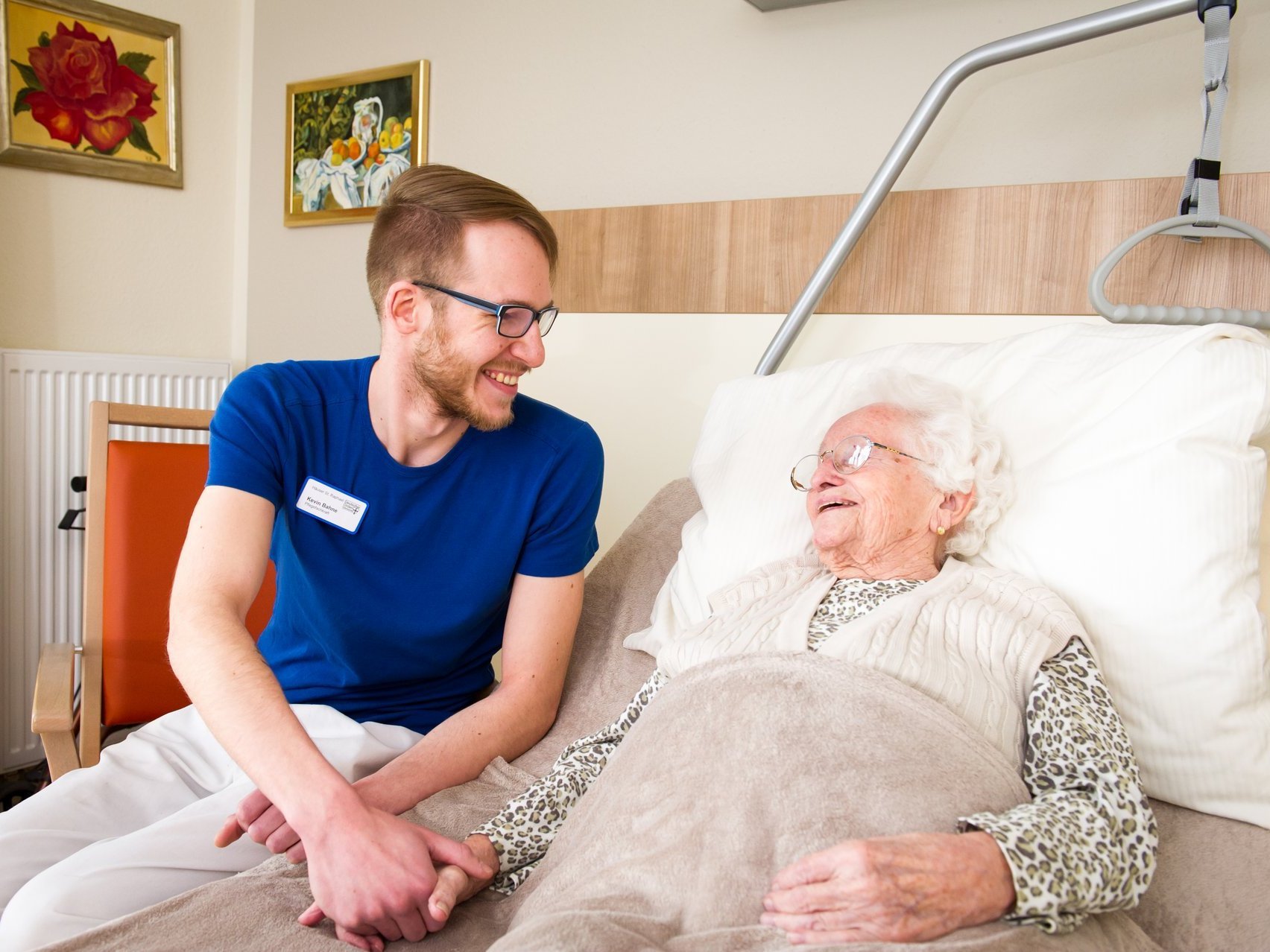 Ein junger Pfleger hält die Hand einer alten Frau, die im Krankenbett liegt - beide lächeln