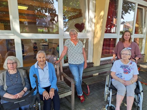 Fünf Frauen posieren vor einem Gebäude mit großen Fenstern. Drei ältere Damen sitzen auf einer Bank und im Rollstuhl. Zwei jüngere Frauen stehen dahinter. Alle blicken freundlich in die Kamera. 