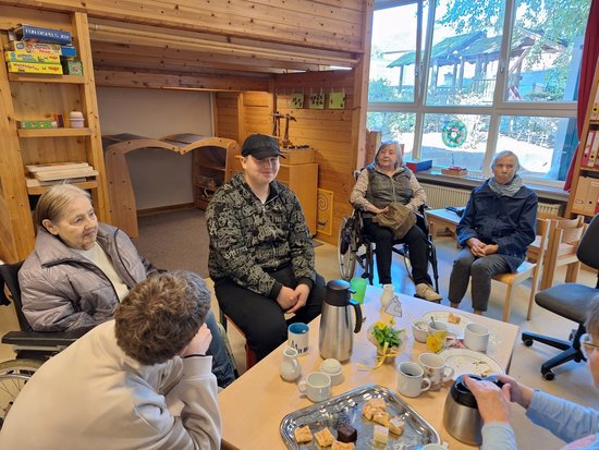 Eine Gruppe älterer Menschen sitzt in einem Raum mit Holzwänden im Kreis zusammen. Zwei Personen befinden sich in Rollstühlen. Auf einem Tisch im Vordergrund stehen Kaffeetassen, eine Thermoskanne und ein Tablett mit Kuchenstücken, was auf eine gemeinsame Kaffeepause hindeutet.