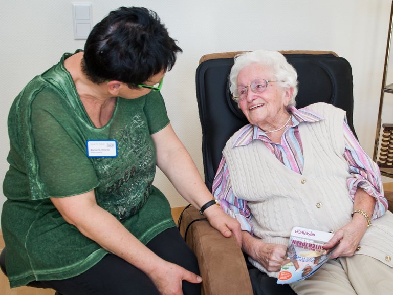 Eine Pflegerin unterhält sich mit einer älteren Frau, die auf einem Massagesessel sitzt